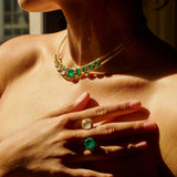 A close-up lifestyle shot of a woman wearing a gold choker necklace with emerald-cut green gemstones and matching emerald ring on her finger, captured in dramatic warm sunlight.