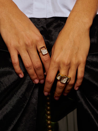 Close-up of a person's hands wearing three gold rings with large clear rectangular gemstones, styled with a white shirt and black satin pants.