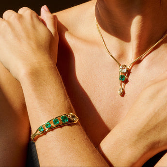 A woman in profile wearing gold jewelry, including a gold necklace with a green pendant, a gold bracelet with green stones, and matching drop earrings, with rings on her fingers. She is photographed in dramatic, warm sunlight with dark shadows.