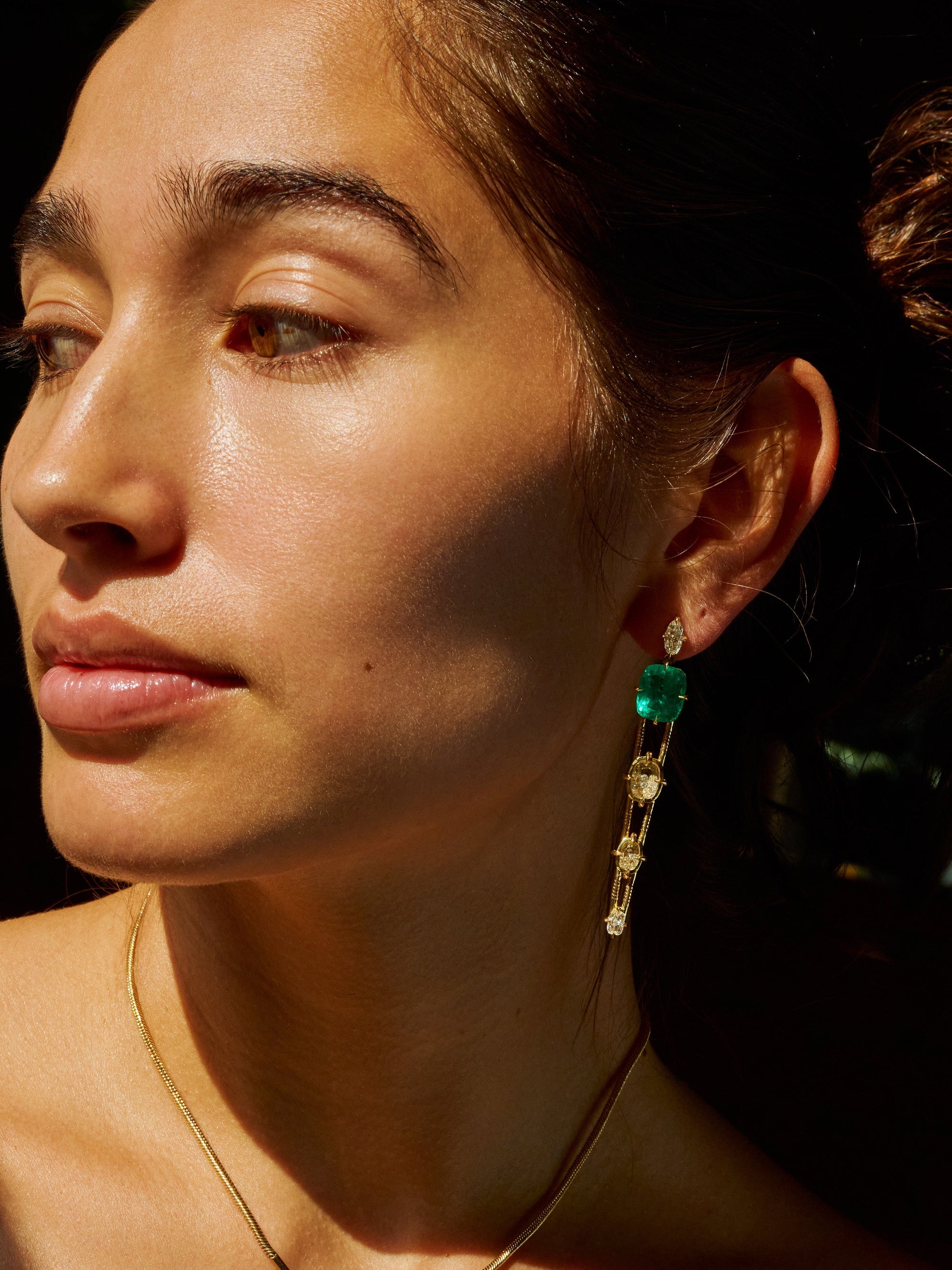 Close-up portrait of a woman wearing a gold necklace with a green and clear stone pendant and a matching drop earring, set against a dark background with dramatic sunlight and shadows.