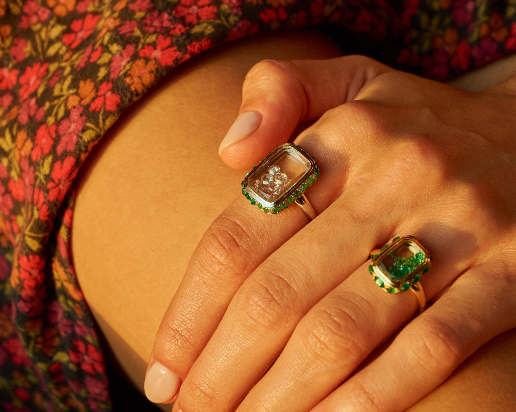 Close-up of a hand wearing two emerald shaker rings on a floral-patterned fabric background.