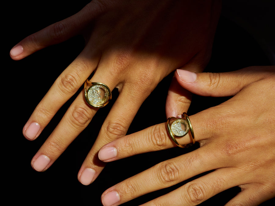 Two hands wearing gold signet rings with oval clear stone settings, posed against a solid black background.